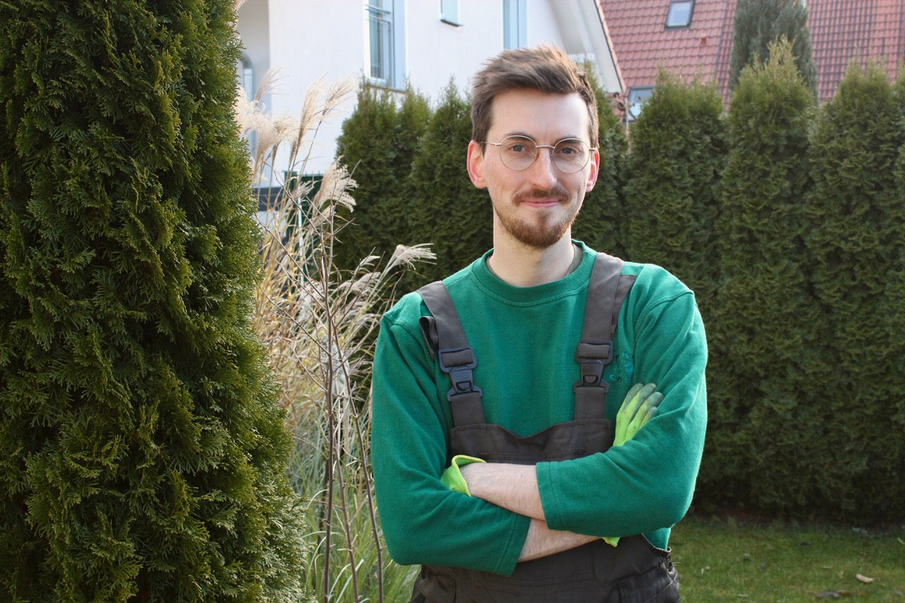 Person in Arbeitskleidung steht im Garten mit verschränkten Armen